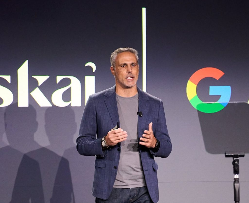 Un homme vêtu d'un t-shirt gris et d'un blazer bleu s'exprime au micro sur scène, debout devant un écran sur lequel s'affichent un extrait de texte et le logo coloré de Google G.