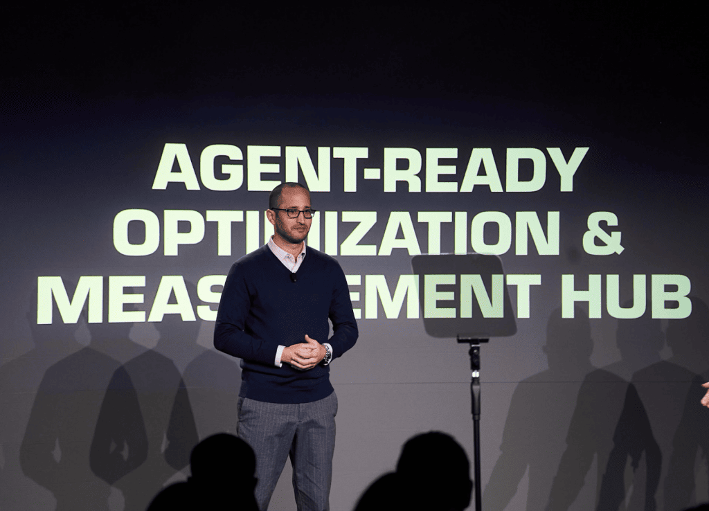 Un homme portant des lunettes et un pull bleu marine se tient sur scène devant un écran sur lequel s'affiche l'inscription « Agent-Ready Optimization & Measurement Hub ». On aperçoit les ombres du public au premier plan.