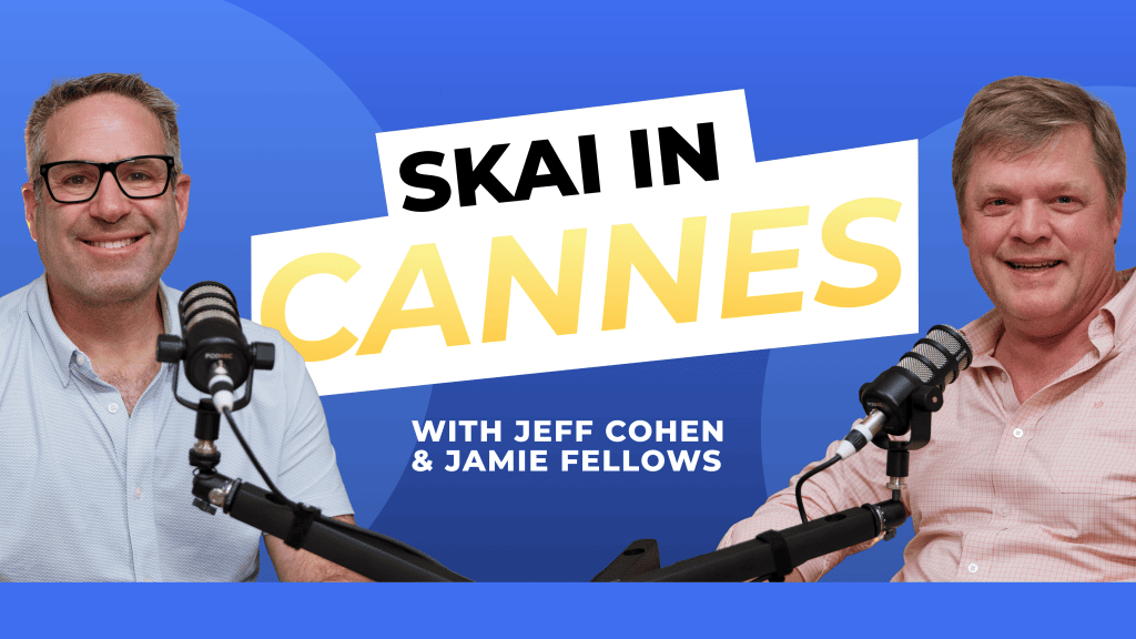 Zwei Männer sitzen lächelnd vor Mikrofonen. Hinter ihnen steht auf blauem Hintergrund mit grafischen Formen der Schriftzug SKAI CANNES“ mit Jeff Cohen & Jamie Fellows.