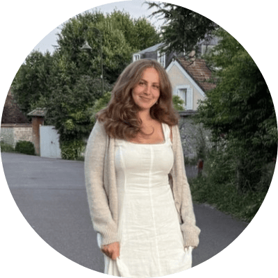 Eine Frau mit langen, welligen braunen Haaren, die ein weißes Kleid und eine beige Strickjacke trägt, steht in einer ruhigen Straße mit Bäumen und Häusern im Hintergrund und verkörpert damit das Wesen einer Retail-Media-Fallstudie in einer beschaulichen Vorstadtkulisse.